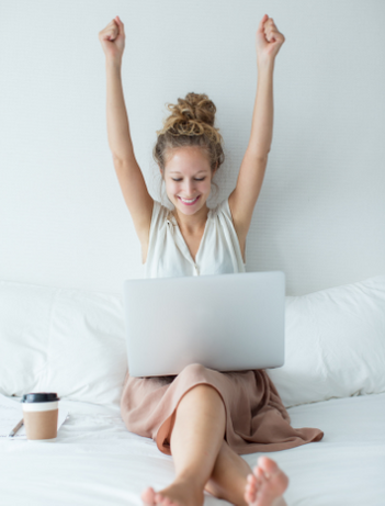 Caucasian woman with arms lifted up in joy while looking at her laptop. There is a cup of coffee to her right.