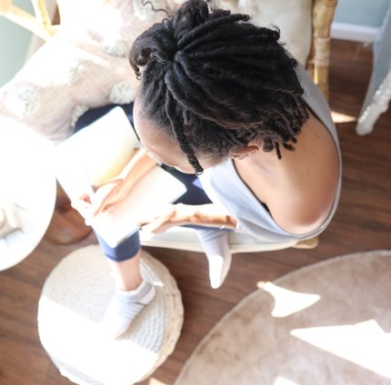 A Black Woman with locs, writing in a notebook. She is seated on a chair with a plush rug in front of her and the sunlight is spilling into the room she's in.